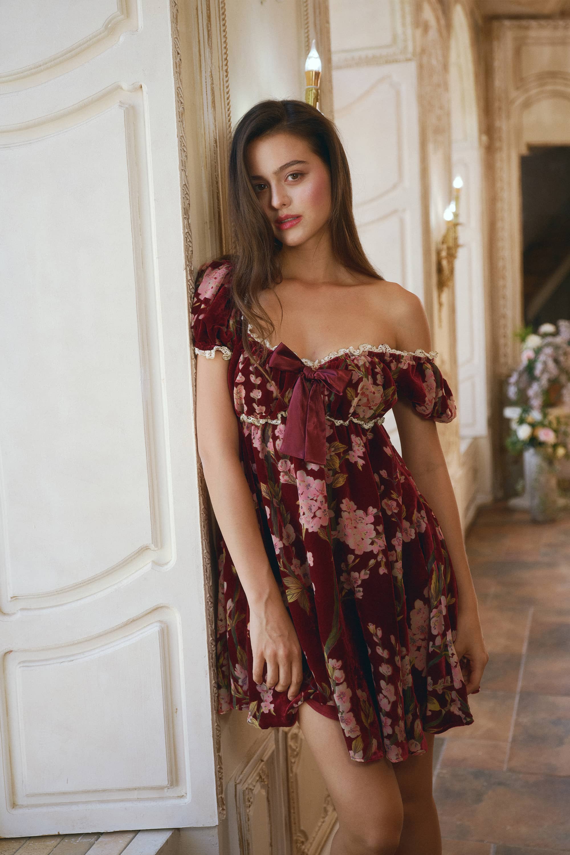 A woman models an off-the-shoulder floral velvet dress in a luxurious interior hallway with cream-colored walls and gold sconces.