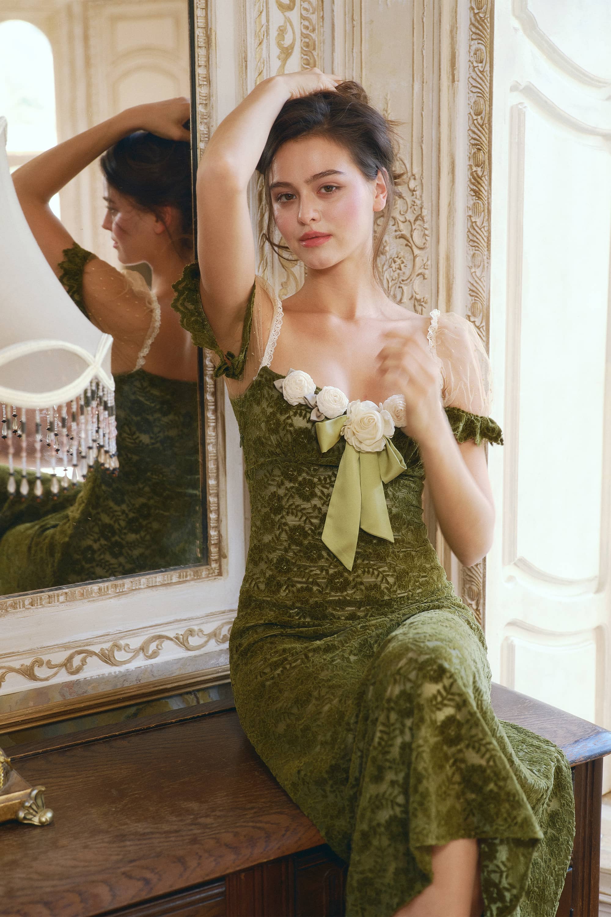 A woman wearing an olive green velvet dress with white floral accents and a large green bow sits on a wooden bench in front of an ornate mirror, with her reflection visible.