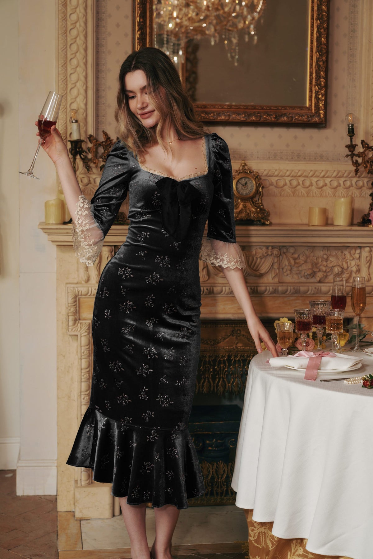 A woman models a black velvet midi dress with floral embroidery and lace flutter sleeves in an elegant, vintage-style room with a fireplace and chandelier.