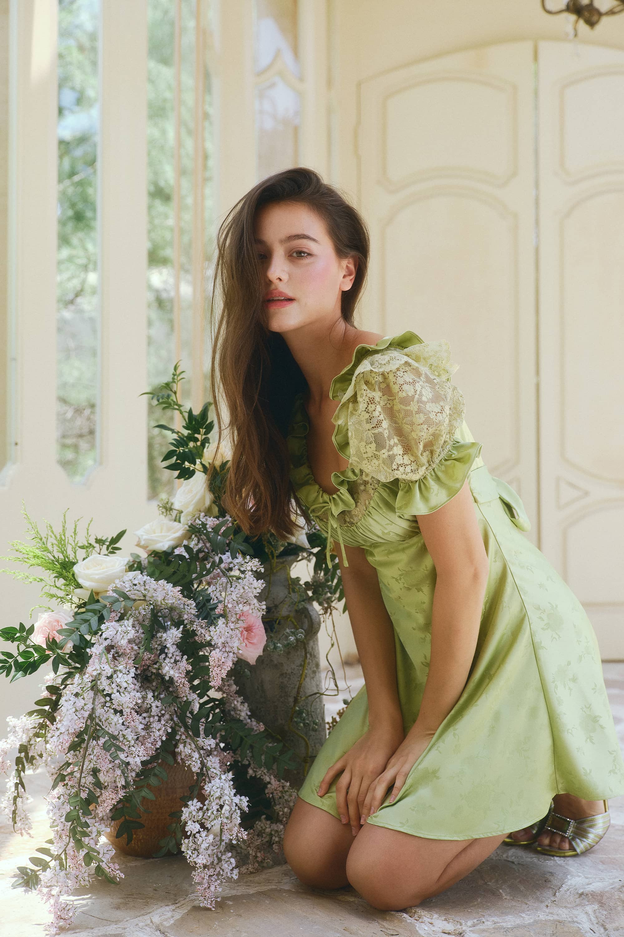 A woman kneels beside a large floral arrangement in a bright, elegant interior. She is wearing a light green dress with ruffled sleeves and lace detailing.
