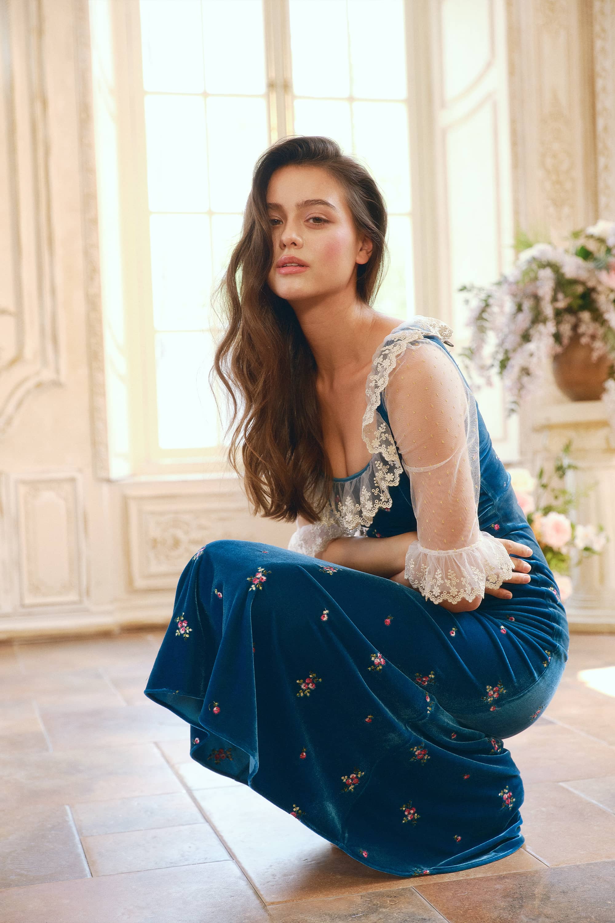 A woman crouches in a sunlit interior wearing a teal-blue velvet dress with floral embroidery and sheer lace sleeves.