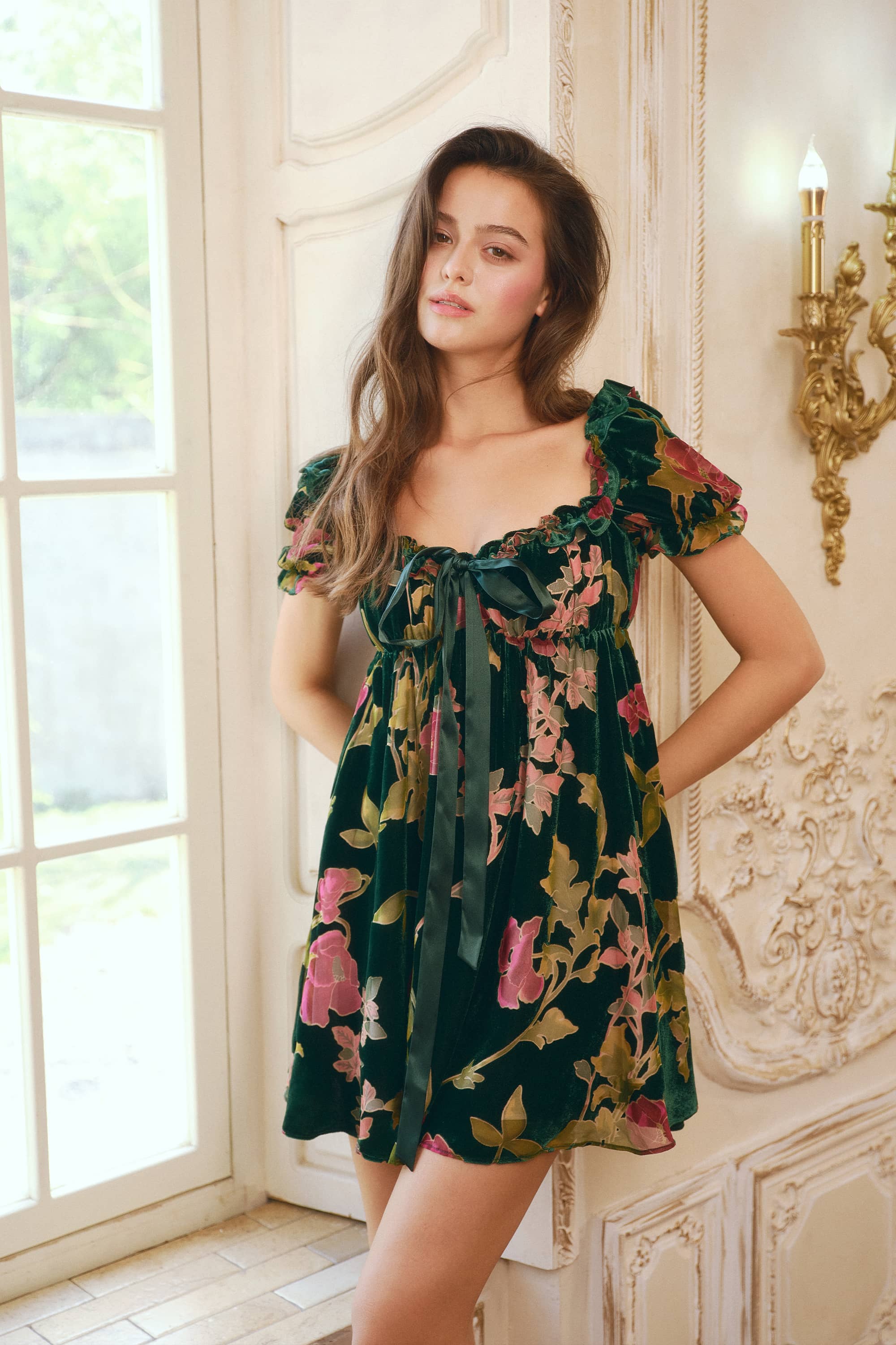 A woman wearing a dark green floral mini dress with puff sleeves and a tie neckline, standing in a classic cream-colored room with ornate wall details and a window.