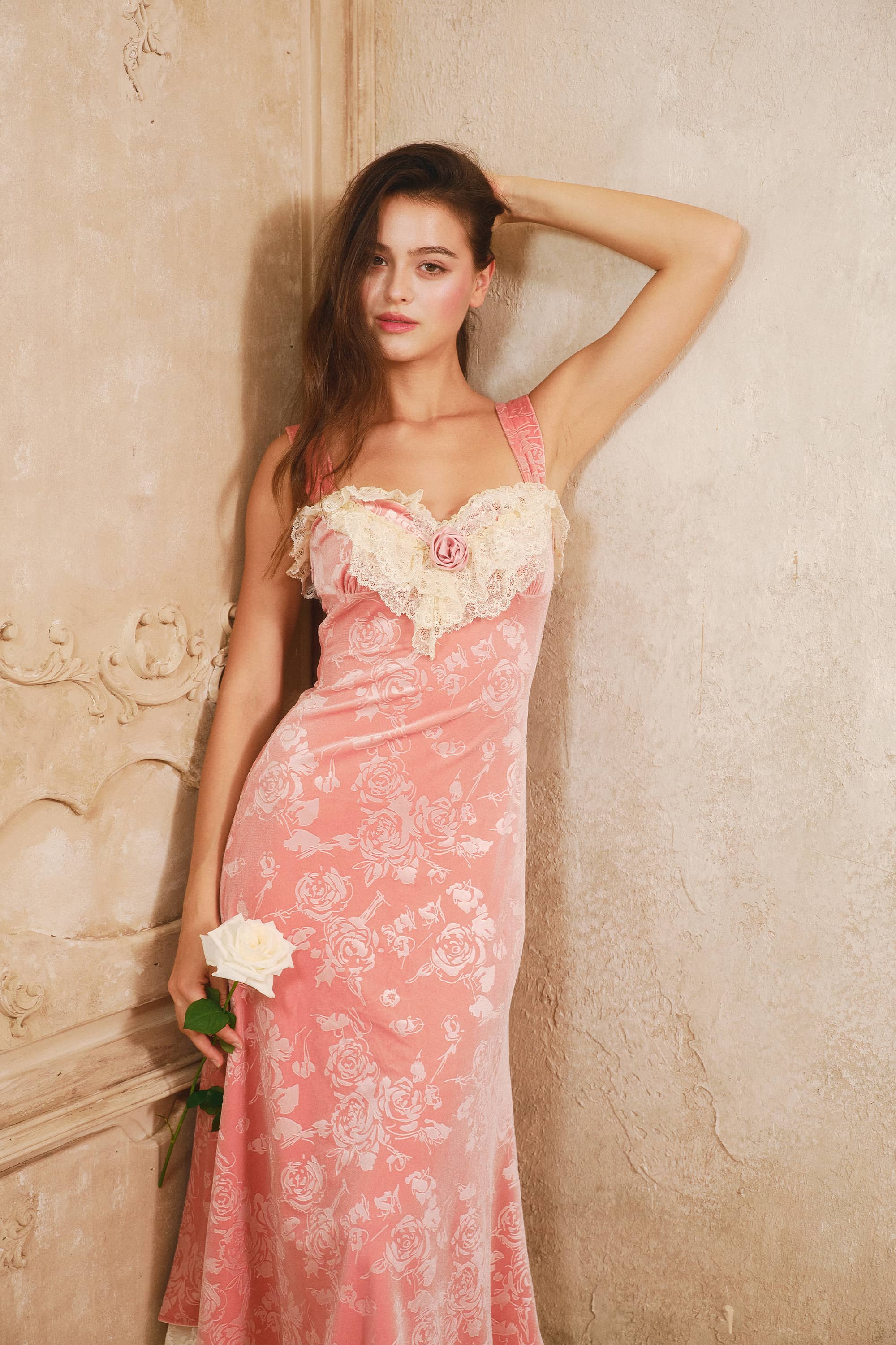A woman models a pink floralvelvet slip dress with a sweetheart neckline and cream lace trim, posing against a textured beige wall.