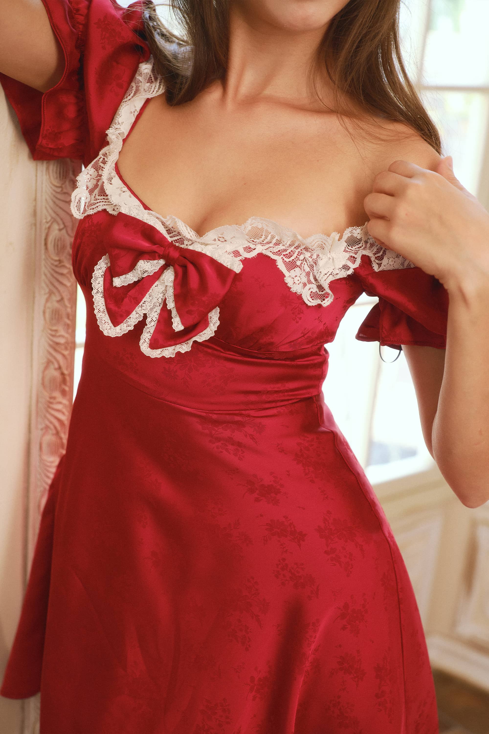 A close-up of a woman wearing a red satin dress with white lace trim and a large bow on the chest, posed indoors near a window.